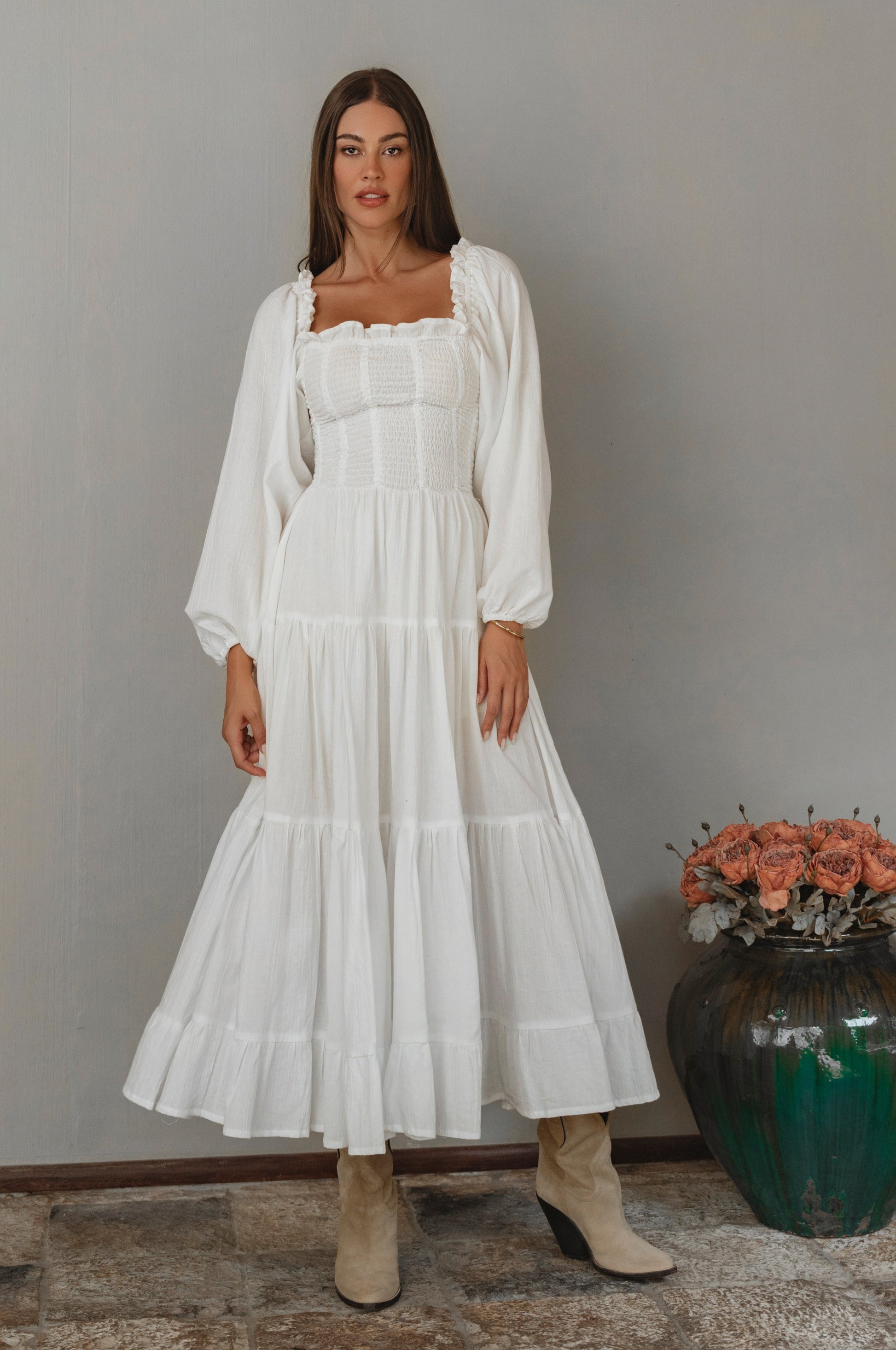 Woman wearing a white long sleeve dress standing next to a vase with flowers against a gray wall.