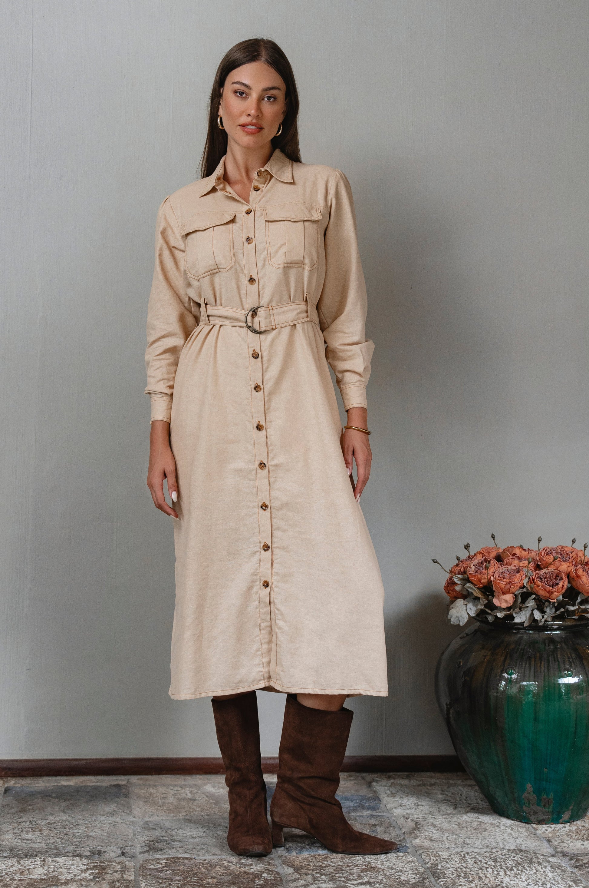 Woman wearing a beige dress with buttons standing next to a vase with flowers against a plain wall.