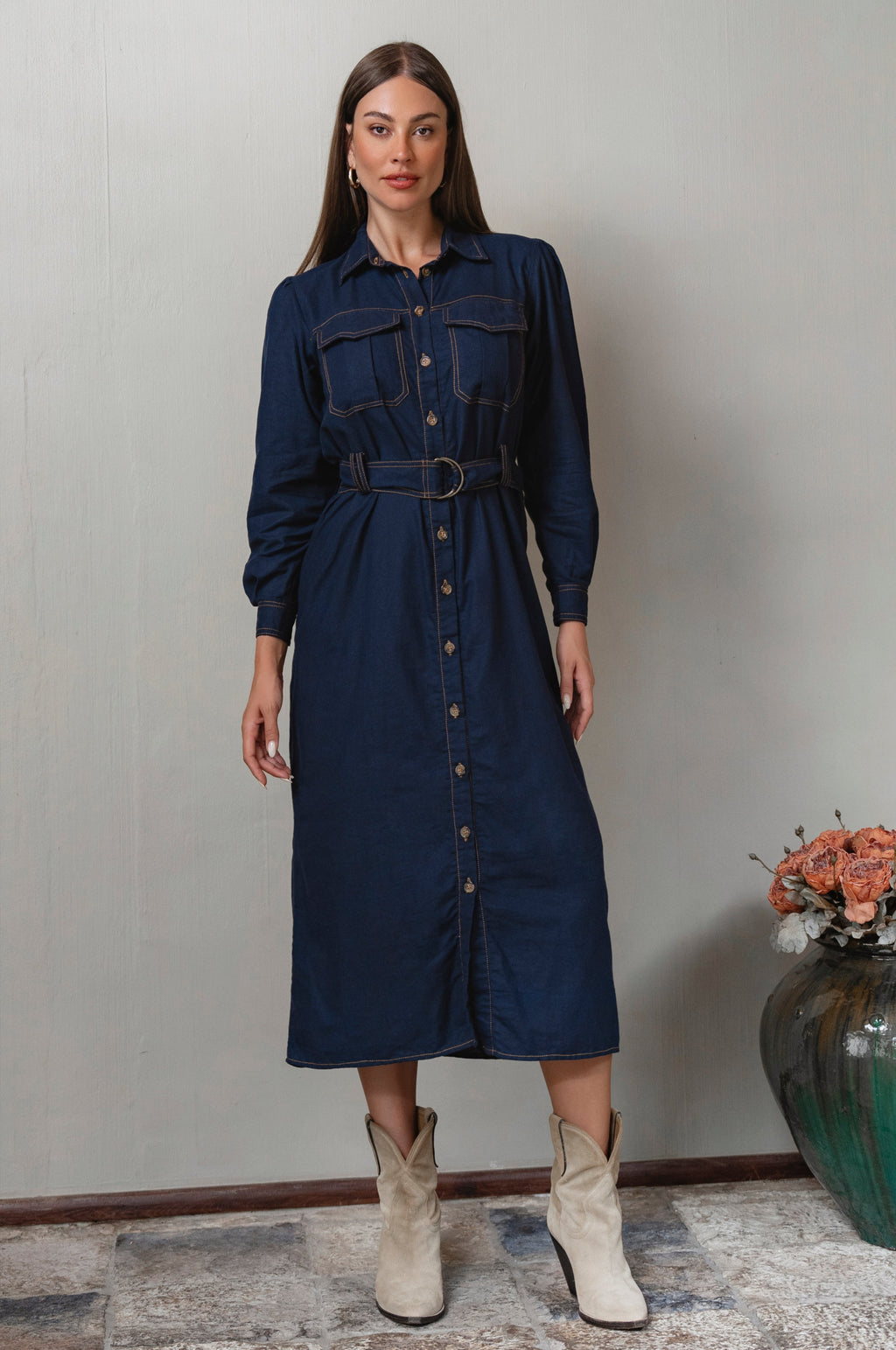 Woman wearing a denim dress with a belt, standing against a plain wall.